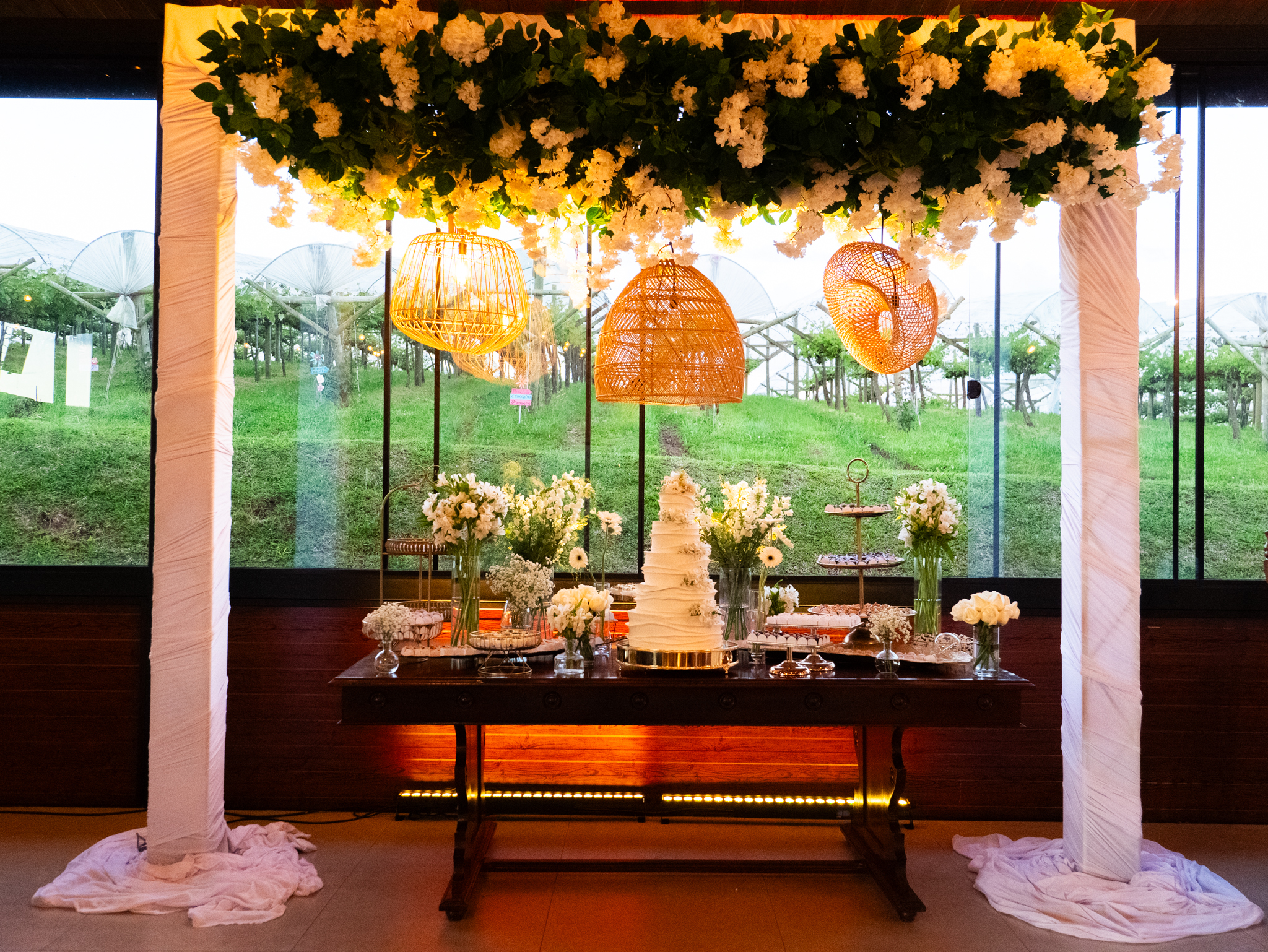 Mesa de doces sob arco de flores brancas e luminárias de palha, com o vinhedo ao fundo
