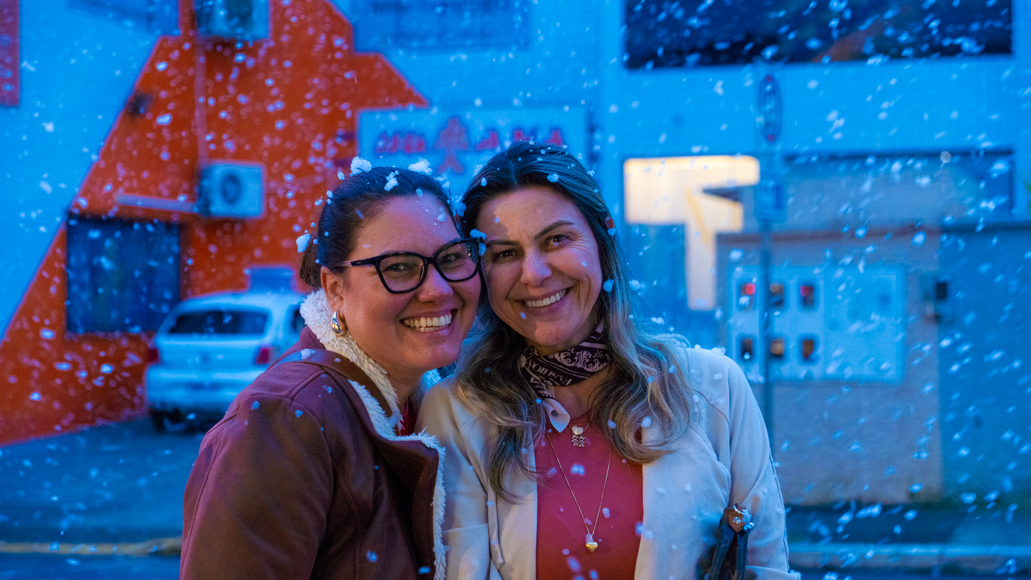 Duas amigas sorrindo, rosto colado, com flocos brancos caindo