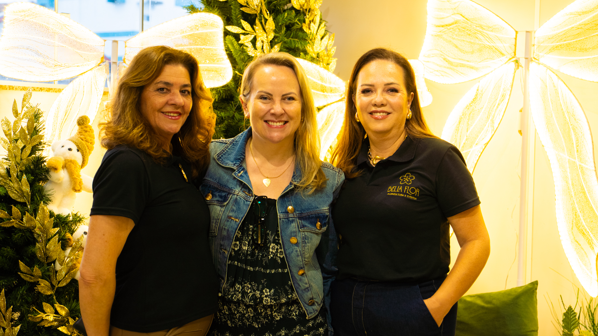 Três mulheres sorrindo em frente à decoração de Natal com laços iluminados