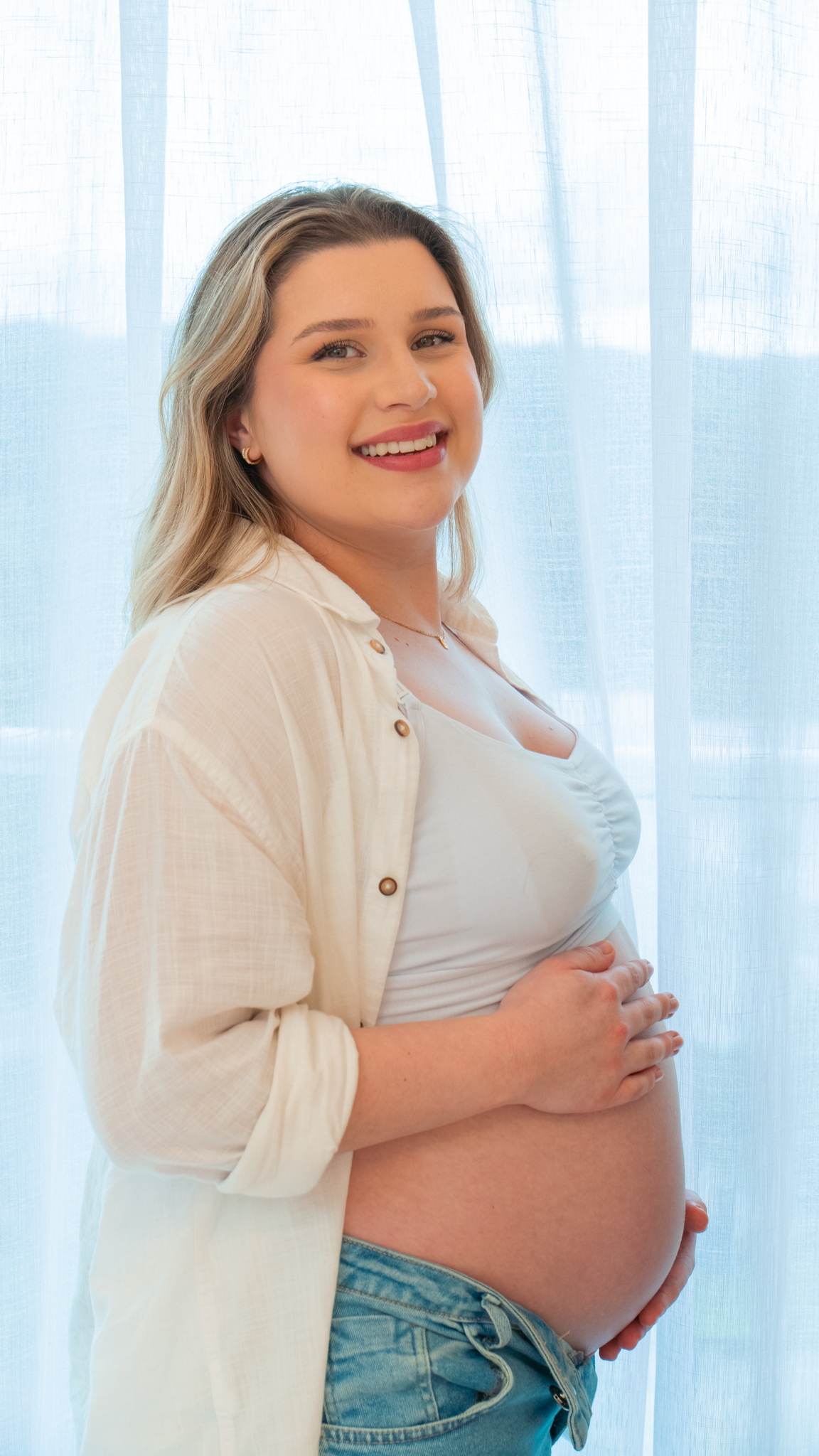 Gestante sorrindo para a câmera, com as mãos na barriga, em frente à cortina