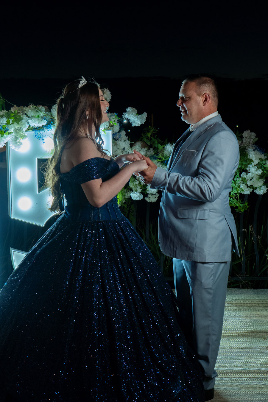 Debutante de mãos dadas com o pai, sorrindo, à noite