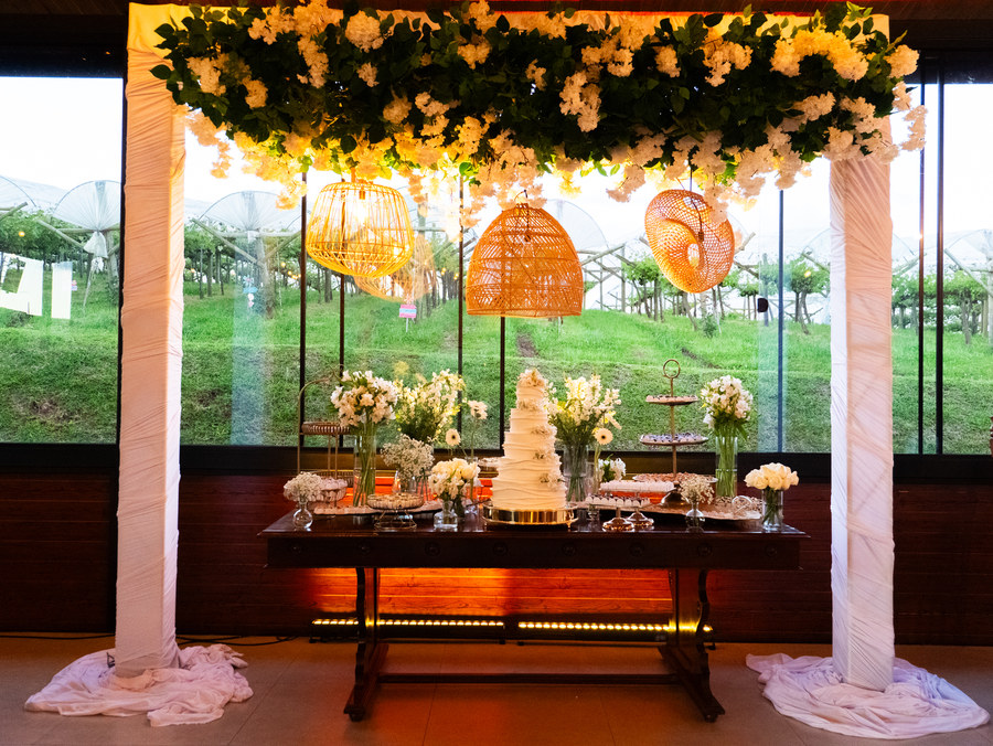 Mesa de doces sob arco de flores brancas e luminárias de palha, com o vinhedo ao fundo