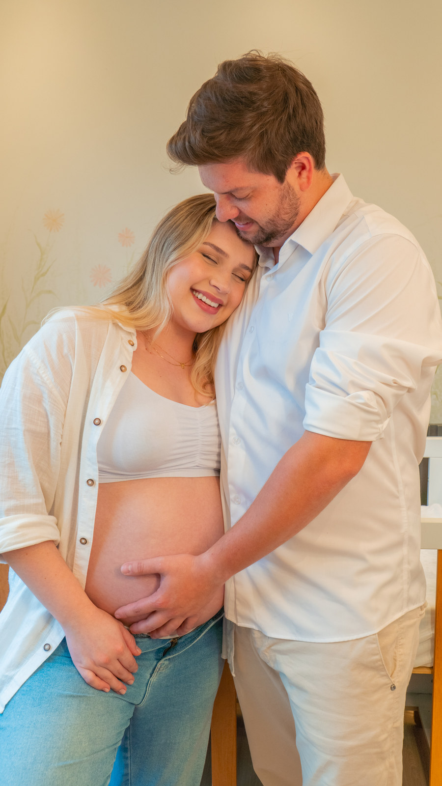 Gestante encostada no ombro do marido, que segura a barriga, no quarto do bebê