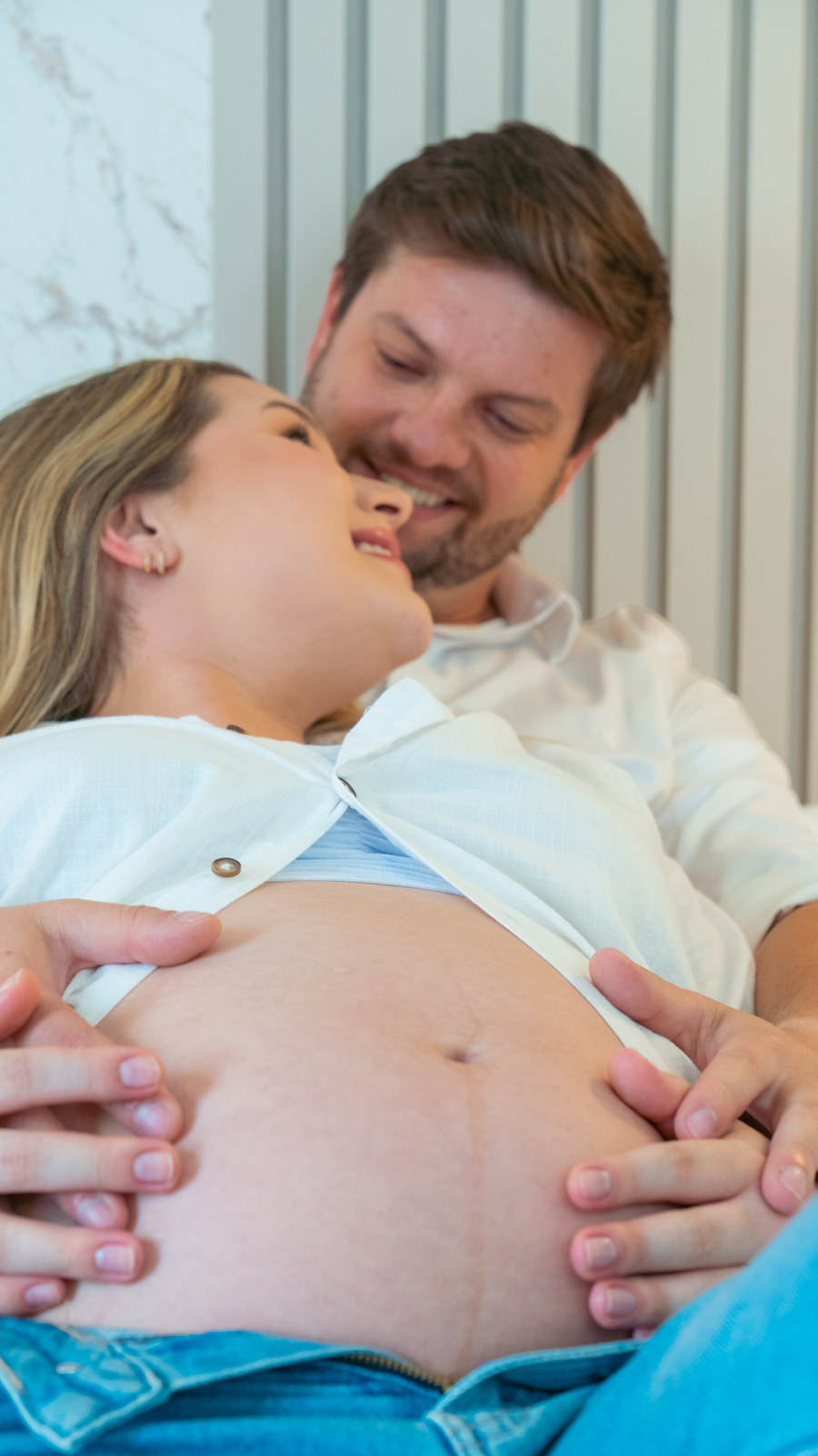 Gestante recostada no marido, os dois sorrindo, com as mãos na barriga