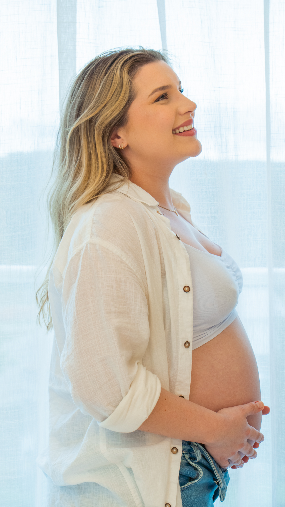 Gestante sorrindo de perfil, com as mãos na barriga, em frente a uma cortina clara